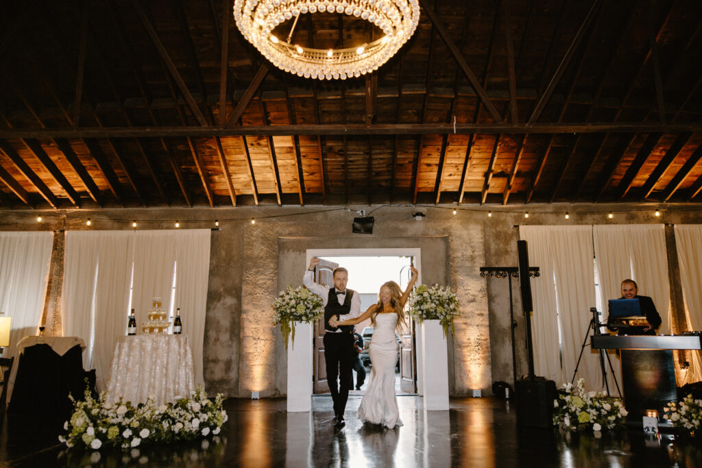 Bride and groom entering their wedding reception with arms raised, dancing under a chandelier in a rustic venue