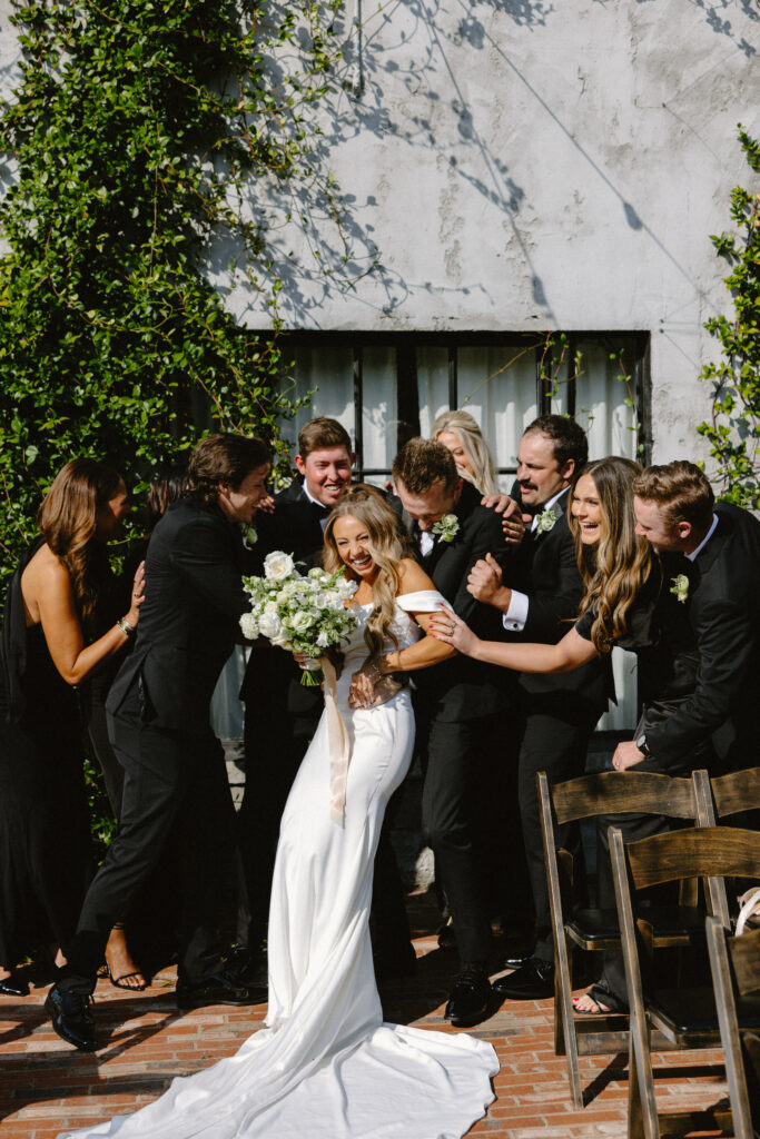 Bride and groom surrounded by their wedding party laughing and hugging in bright outdoor sunlight