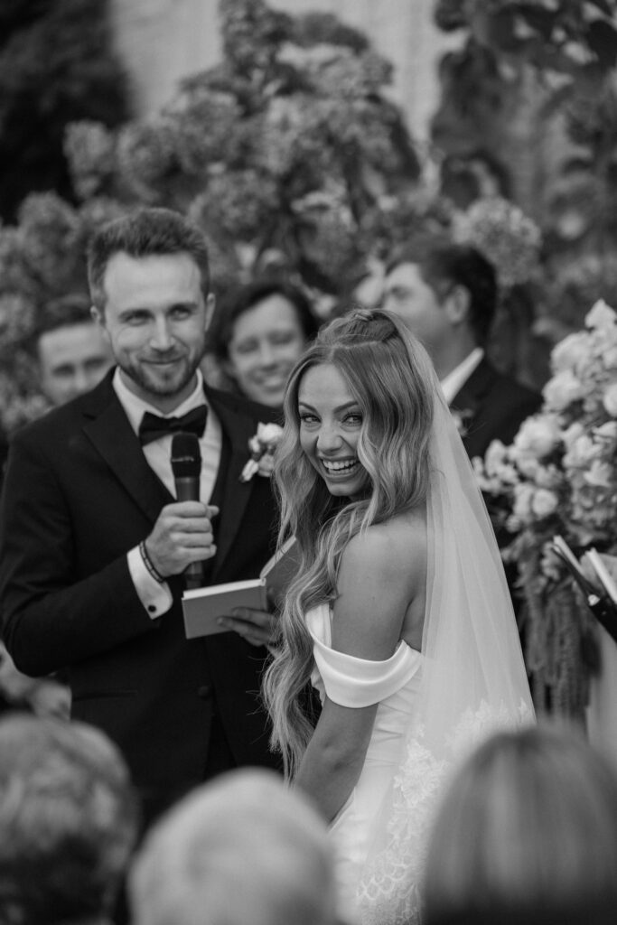 Bride smiling back at guests during ceremony while groom reads vows at the altar