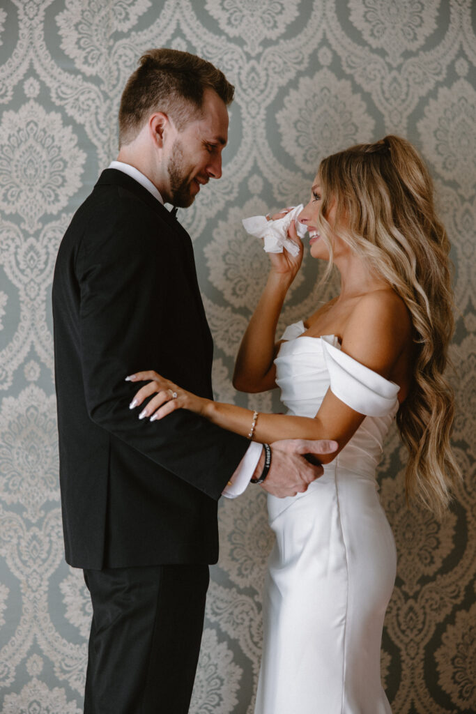Bride wiping away tears while smiling at groom during their emotional first look on wedding day
