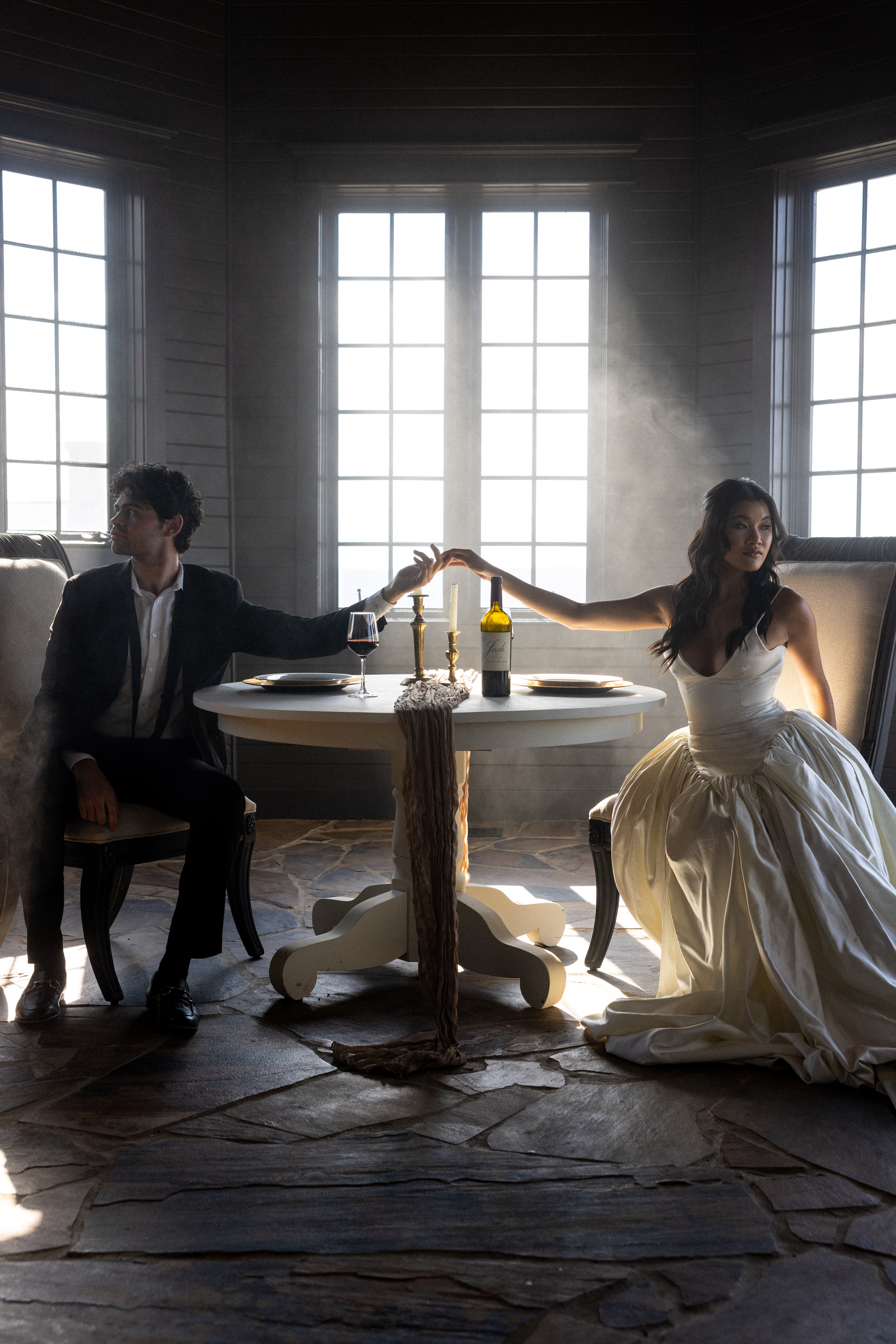 Couple sitting on opposite sides of a round table in dramatic window light, reaching toward each other through soft smoke with wine and candles between them.