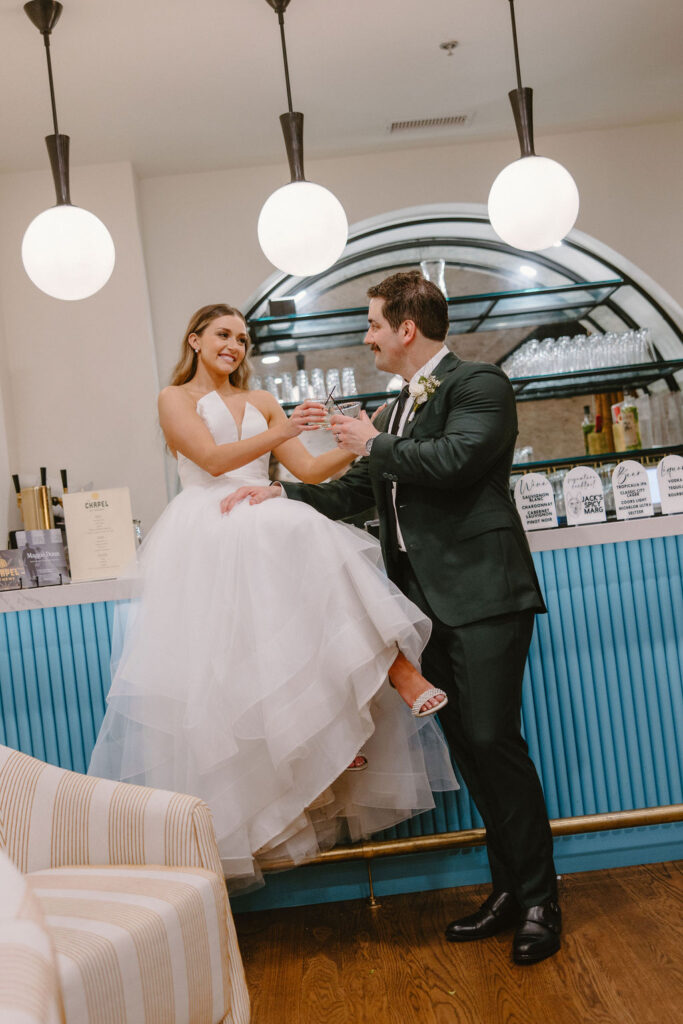 Bride and groom sitting at the bar cheering their signature wedding cocktails while looking into each others eyes