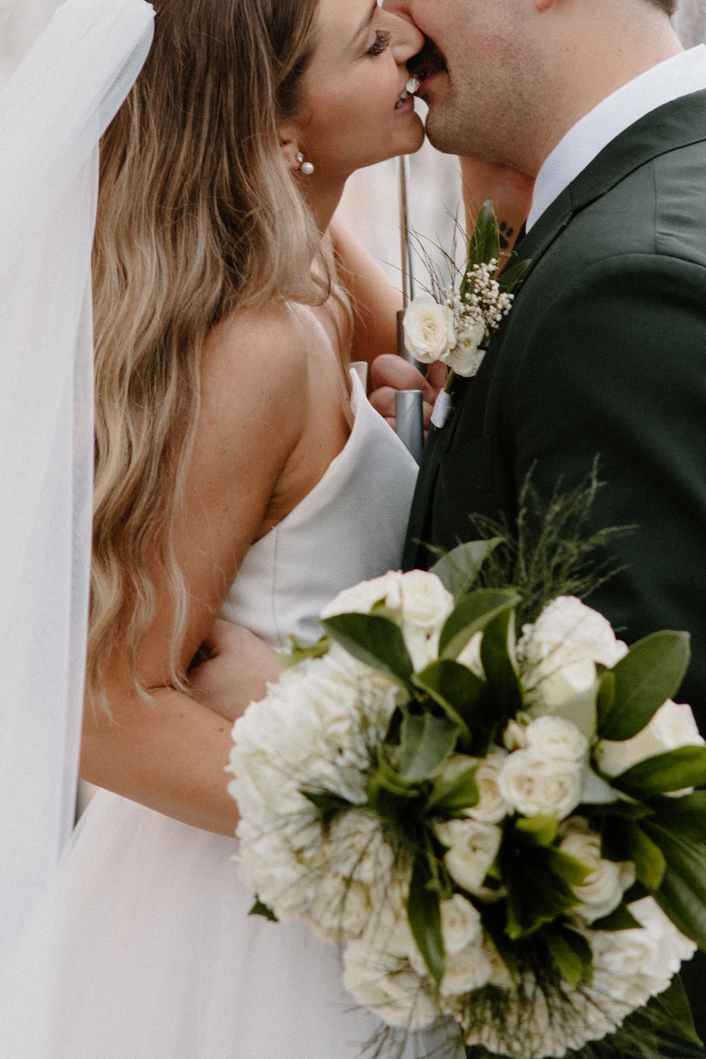 Bride and Groom in a dark green suit under a clear umbrella kissing in the rain during their wedding couples portraits