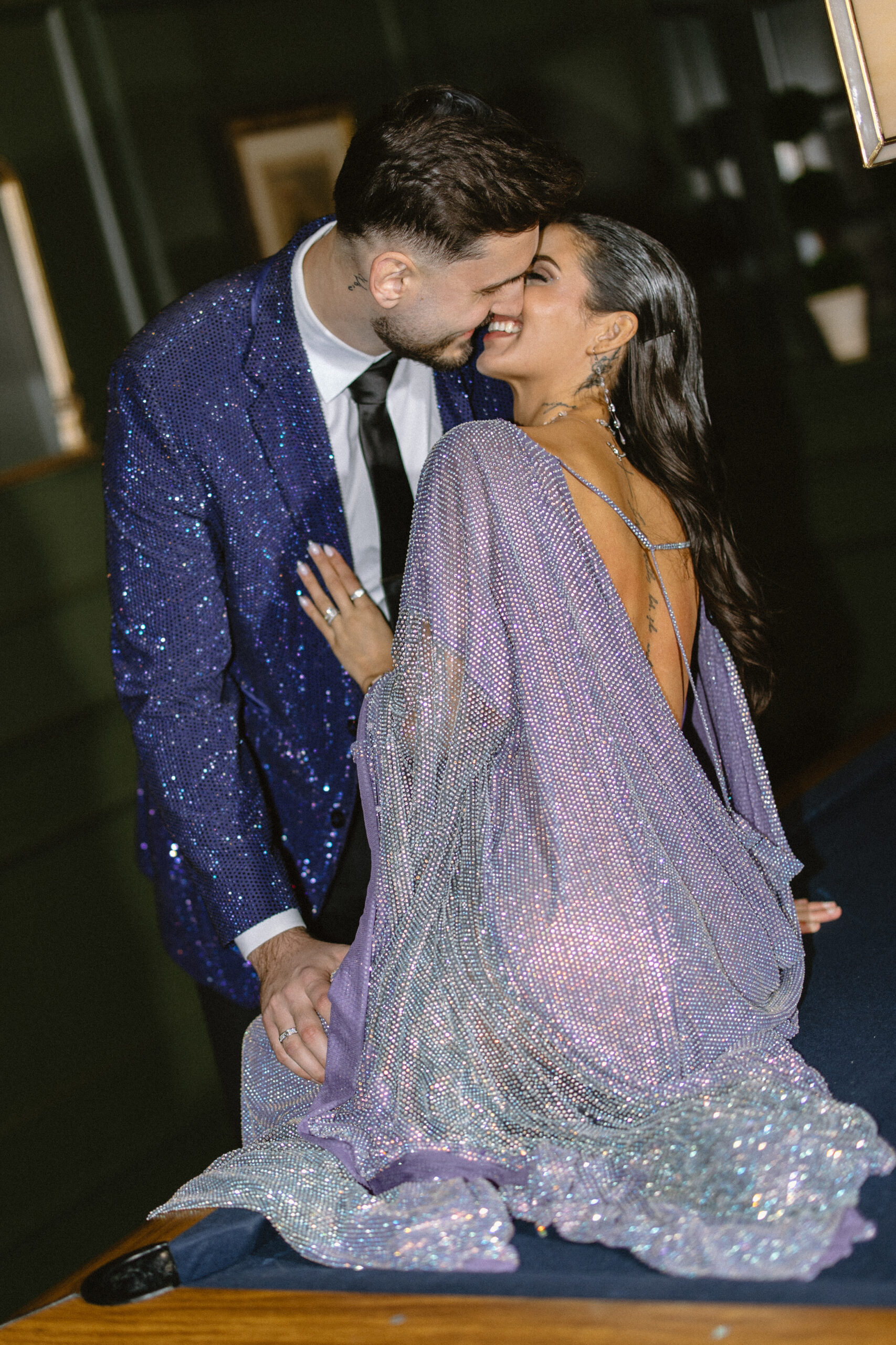 Groom in blue sequin suit kissing bride wearing a translucent draped purple dress on pool table in the lounge area during their wedding reception