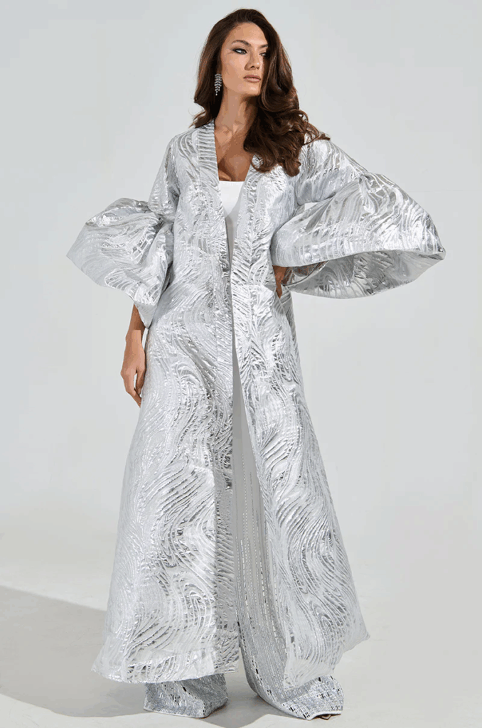 Person wearing a long metallic silver coat with oversized sculptural sleeves and textured fabric, styled with wide leg matching pants for an editorial engagement look.