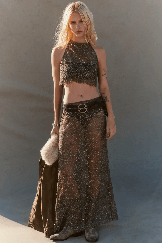 Person wearing a sheer sequin maxi skirt with matching cropped halter top, styled with a double belt and worn boots in warm natural light.