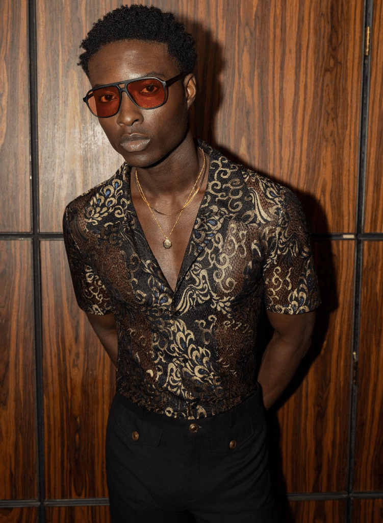 Person wearing a sheer black and gold brocade short sleeve shirt paired with tailored black trousers and red tinted sunglasses for a bold fashion forward engagement look.