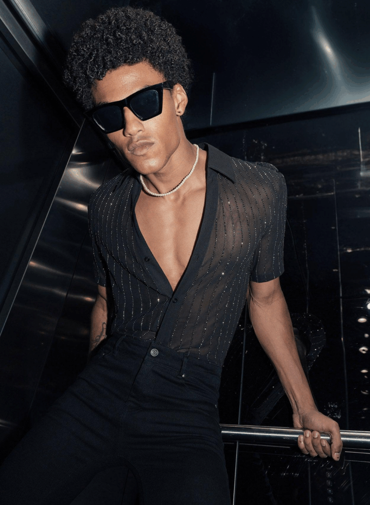 Person wearing a sheer black short sleeve button down shirt with vertical crystal embellishments, styled with high waisted black trousers and dark sunglasses.