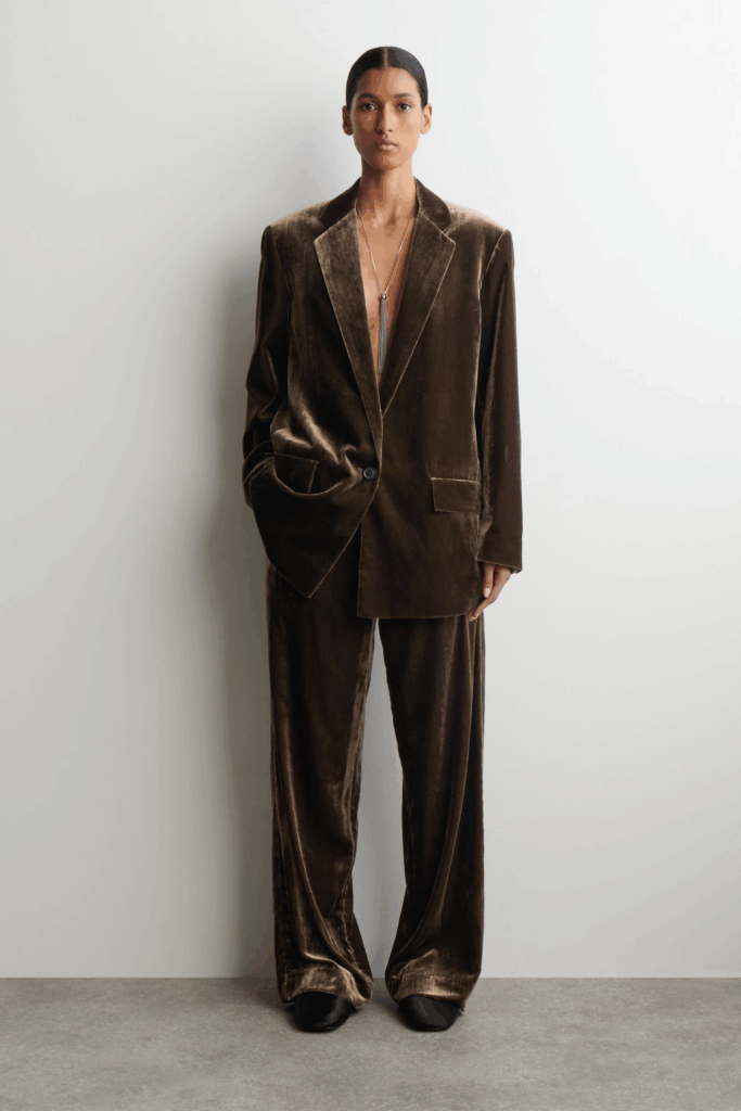 Person wearing a chocolate brown velvet oversized blazer and matching wide leg trousers styled with minimal jewelry for an elevated genderless engagement outfit.