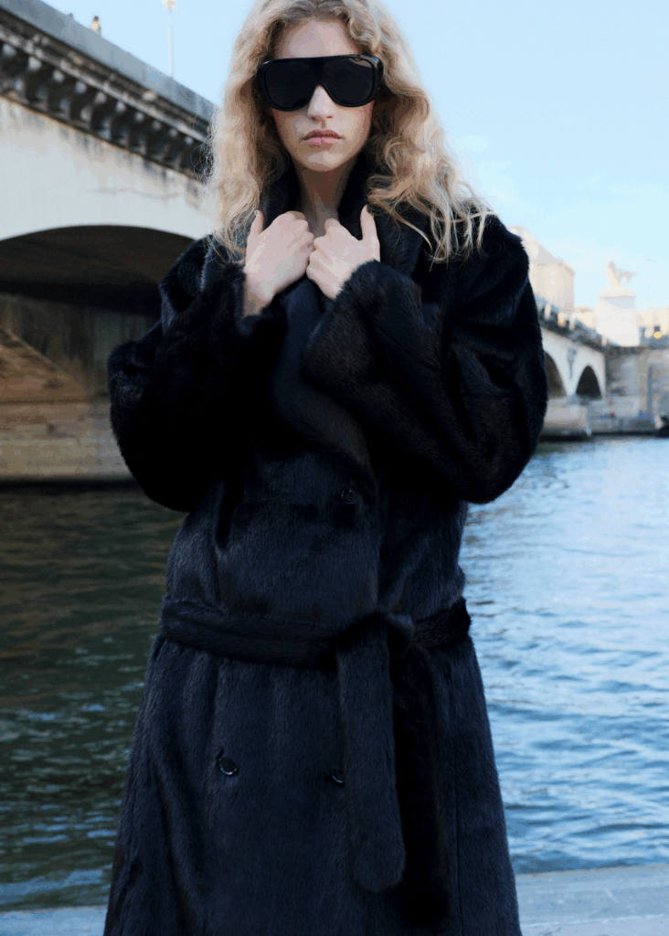 Person wearing an oversized black faux fur coat and dark sunglasses standing by the riverside with a Parisian bridge in the background.