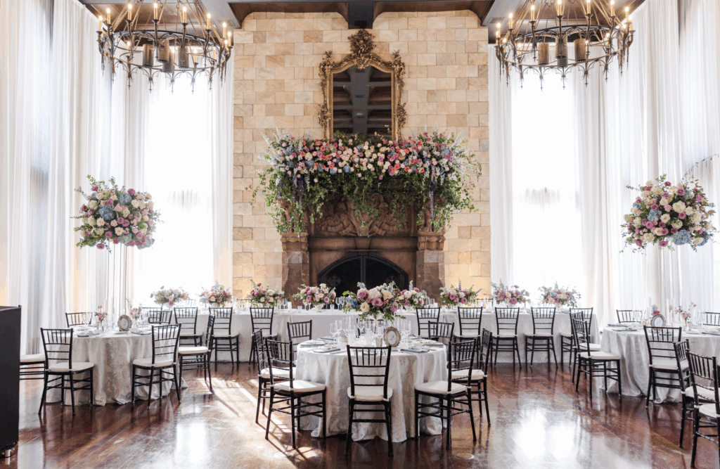 Elegant wedding reception at Dover Hall in Virginia featuring a stone fireplace, hanging chandeliers, round tables with floral centerpieces, and soft natural light through tall draped windows